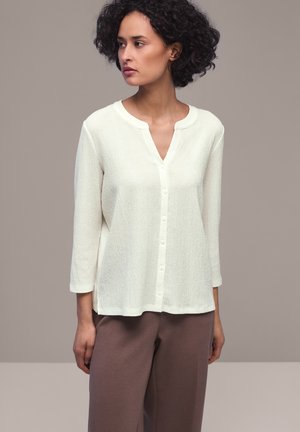 Donna con capelli ricci che indossa una camicetta bianca texturizzata con bottoni e maniche a tre quarti e pantaloni marroni a gamba larga, in piedi davanti a uno sfondo semplice.