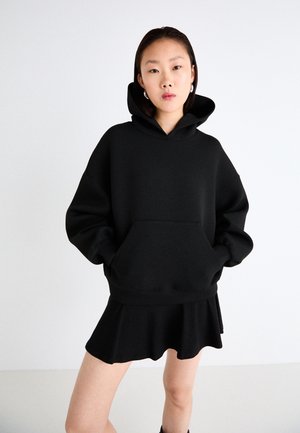 Black oversized hoodie with a front pocket, ribbed cuffs, and hem. Paired with a flared skirt. Smooth fabric texture, minimal design.