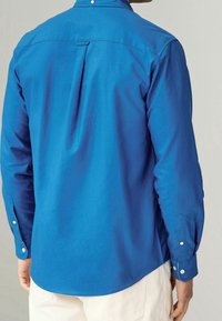 Camisa azul de manga larga con un pliegue en la espalda, cuello abotonado y dobladillo redondeado. El material parece suave, con detalles de botones en los puños.