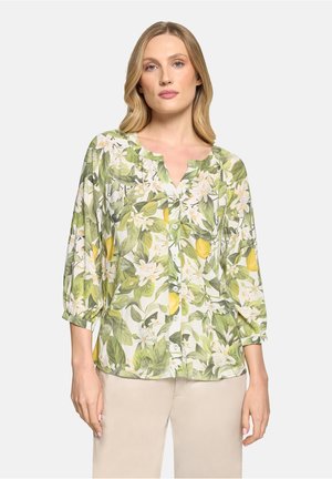 Kvinde iført en knaplukket, blomsterprintet bluse med grønne blade og gule citronmønstre, kombineret med lyse beige bukser.