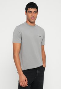 Άνδρας με κοντά σγουρά μαλλιά, φορώντας ένα ανοιχτό γκρι t-shirt BOSS και μαύρα τζιν, στέκεται με το ένα χέρι στην τσέπη μπροστά σε λευκό φόντο.