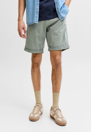 Man staat met één hand in de zak, draagt opgerolde grijze shorts, beige sokken, beige sneakers, een donkerblauw overhemd en een lichtblauw spijkerhemd.