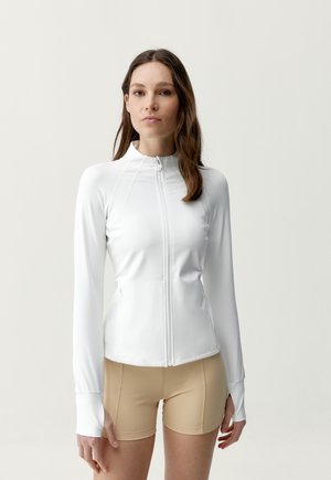 Žena oblečená v priliehavom bielom zipsom na zapínanie na dlhý rukáv a béžových cyklistických šortkách, stojaca pred jednoduchým svetlým pozadím.