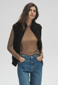 Brun långärmad topp under en svart fleeceväst; modellen bär blå denimjeans med avslappnad passform. Dragkedjedetaljer på västen.