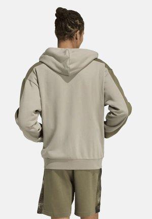 Personne aux cheveux tressés portant un sweat à capuche gris clair et un short vert olive, vue de dos avec les mains sur les hanches.