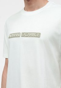 Άνδρας που φοράει λευκό T-shirt με μπεζ κεντημένο κείμενο "Armani Exchange" στο στήθος.
