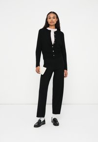 Cardigã preto com fecho de botão, combinado com calças pretas de corte descontraído. O modelo segura uma clutch cremosa texturizada. Os sapatos são mocassins pretos brilhantes.