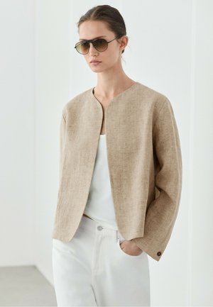 Donna che indossa una giacca di lino beige aperta davanti, top bianco, pantaloni bianchi e occhiali da sole aviator scuri, in piedi con una mano in tasca.