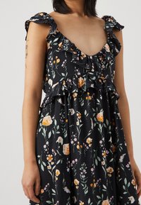 Mustaton musta mekko, jossa on rypytetyt hihnat ja kukkakuvio oranssilla, valkoisella ja vihreällä, jota käyttää henkilö, jolla on näkyvä tatuointi käsivarressaan.