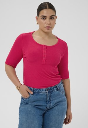 Žena s tamnom kosom u ružičastoj bluzici s gumbima i plavim trapericama, stoji s jednom rukom u džepu na jednobojnoj pozadini.