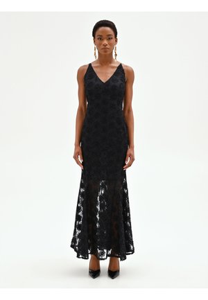 Femme portant une robe noire en dentelle sans manches avec un décolleté en V et une jupe inférieure transparente, associée à des talons pointus noirs et des boucles d'oreilles dorées.