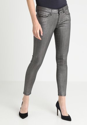 Slim-fit metallic sølvbukser båret med sorte høje hæle og en sort top, vist fra talje til fødder mod en ensfarvet baggrund.