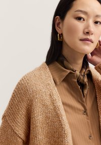 Karamellfarbener Strickcardigan mit weiten Ärmeln, kombiniert mit einem braun gestreiften Hemd und einem braunen Schal. Goldene geometrische Ohrringe setzen Akzente.