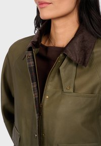 Skórzana kurtka w oliwkowym kolorze z brązowym kołnierzem z kordurowej tkaniny, w kratkę podszewką, złotym zamkiem i przednimi kieszeniami. Gładka tekstura o minimalnym połysku.