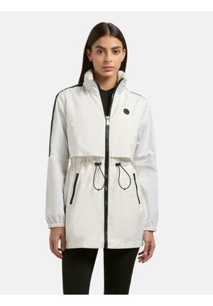 Młoda kobieta ubrana w białą kurtkę na zamek błyskawiczny z czarnymi sznurkami i zamkami, stojąca na tle jednolitego jasnego tła.