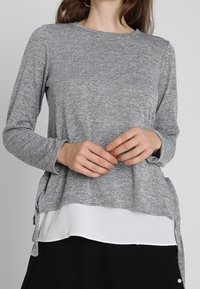 Szary długi rękaw bluzka z okrągłym dekoltem, z warstwowym białym dolnym panelem i bocznymi rozcięciami, wykonana z miękkiego, teksturowanego materiału.