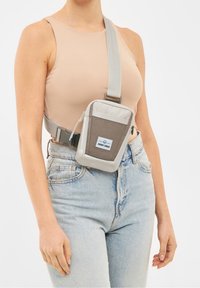 Šedá kabelka přes rameno z tkané látky a hladkého materiálu, s kapsou na zip a nastavitelným popruhem, nošená přes béžový top.