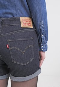 Bermudas de mezclilla, de lavado oscuro, con costuras visibles y dobladillos enrollados. Presenta la etiqueta de Levi Strauss & Co. y un diseño distintivo en el bolsillo trasero.