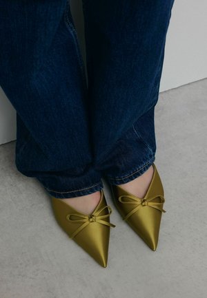 Pointed-toe, metallic yellow-green flats with a satin finish and small bow detail, paired with dark blue jeans. Floor surface is light gray.