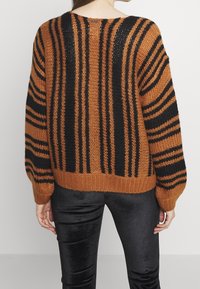 Person, die einen lockeren braun-schwarz gestreiften Strickpullover und schwarze Samt-Hosen trägt, von hinten vor einem schlichten Hintergrund gezeigt.