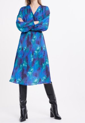 Femme portant une robe midi plissée florale bleue et bleu sarcelle à manches longues et des bottes noires à talons montant aux genoux, bras croisés.