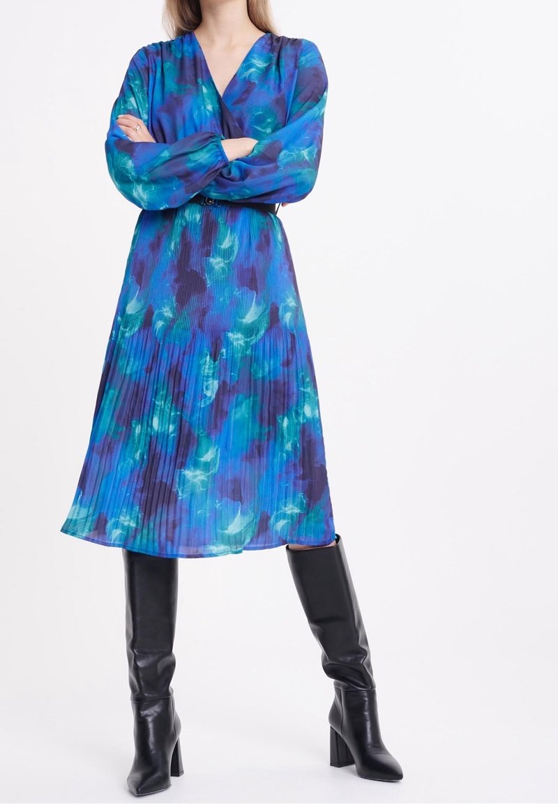 Femme portant une robe midi plissée florale bleue et bleu sarcelle à manches longues et des bottes noires à talons montant aux genoux, bras croisés.
