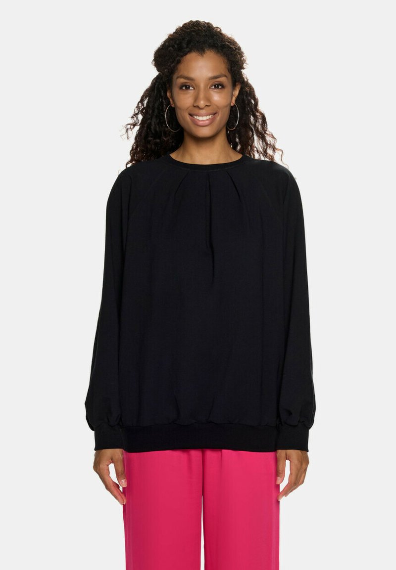 Czarny długi sweter z rękawami, z okrągłym dekoltem, plisowanym przodem i elastycznym dołem. Noszony z jaskraworóżowymi szerokimi spodniami.