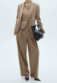 Personne portant un costume beige trois pièces avec un pantalon à jambes larges, tenant un sac à main en cuir noir et portant des talons noirs pointus.