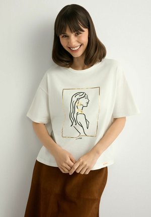 Uśmiechnięta kobieta z włosami do ramion, ubrana w białą koszulkę z czarno-złotym rysunkiem kobiety w profilu oraz brązową spódnicę.