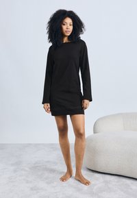 Ženska s kodrastimi lasmi, bosa, oblečena v črno dolgo obleko z dolgimi rokavi, stoji poleg belega teksturiranega fotelja na svetlem preprogi.