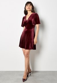 Bordeaux sametové šaty s krátkými nabíranými rukávy, hlubokým V výstřihem, přiléhavým korzetem a rozšířenou sukňí. Nosí se s lodyhovými podpatky.