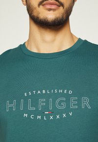Teal t-paita, jossa on pyöreä kaula-aukko ja näyttävä valkoinen graafinen teksti "ESTABLISHED HILFIGER MCMLXXXV" punaisilla ja valkoisilla yksityiskohdilla.