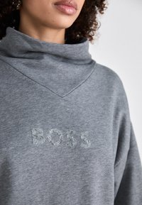 Grauer Kapuzenpullover mit hohem Kragen, der das Wort "BOSS" in Pailletten zeigt. Weicher Stoff und lockere Passform. V-Ausschnitt-Detail.