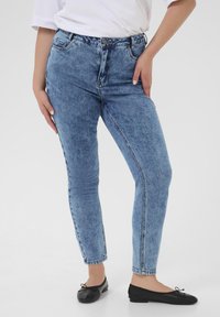 Vaqueros de mezclilla azul con cintura alta, ajuste skinny y acabado descolorido. Cuenta con un cierre de botón y cinco bolsillos. Se llevan con zapatos planos negros.