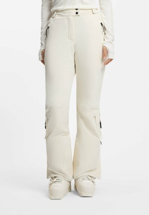 Kremowe ocieplane spodnie narciarskie z bocznymi kieszeniami na suwak i rozszerzanymi nogawkami na dopasowane buty narciarskie, noszone przez stojącą osobę.