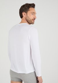 Chemise blanche à manches longues en tissu doux, avec col rond et coupe décontractée, accompagnée de shorts gris.