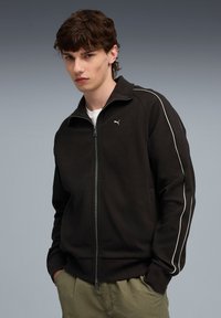 Zwarte rits-sweater van een gestructureerde stof, met witte zijstrepen en een klein logo op de borst. Gecombineerd met lichtgroene broek.