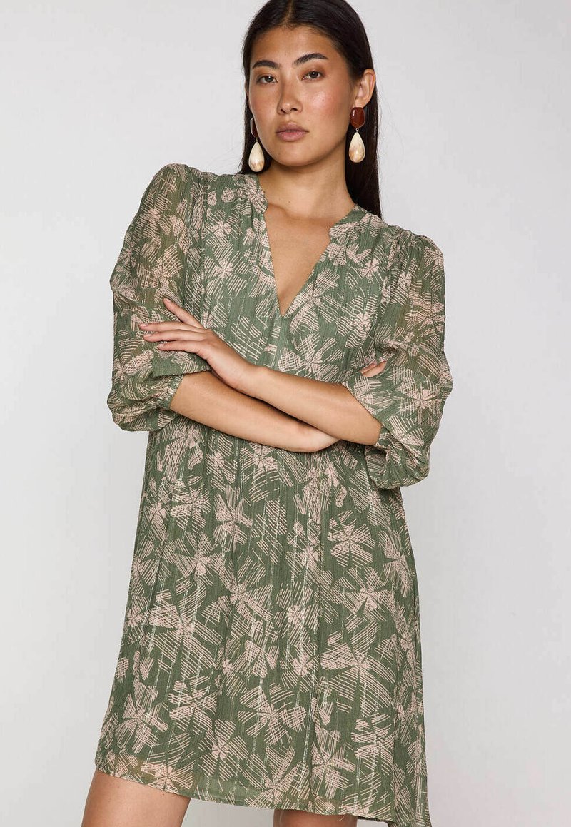 Vrouw met lang donker haar draagt een groene jurk met lichtroze abstracte bloemenprint en grote oorbellen, met gekruiste armen, op een neutrale achtergrond.