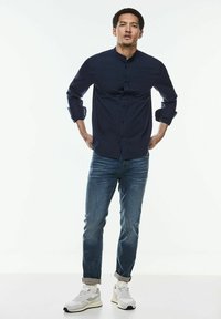 Camisa de botones azul marino con cuello, combinada con jeans de mezclilla azul claro y zapatillas blancas. La camisa tiene mangas largas remangadas en los puños.