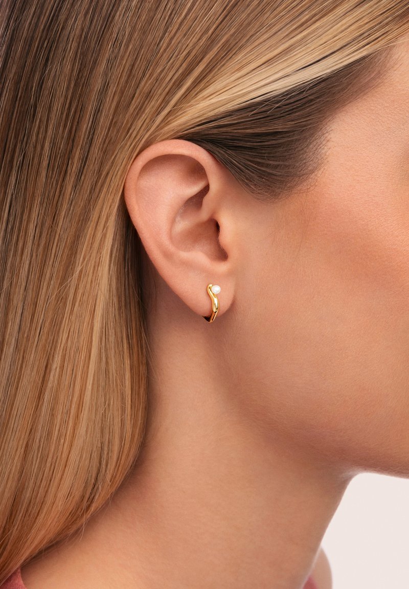 Orecchini a cerchio in oro con un design ondulato, caratterizzati da un piccolo accento di perla. Gli orecchini sono indossati da un modello, evidenziando la loro forma.