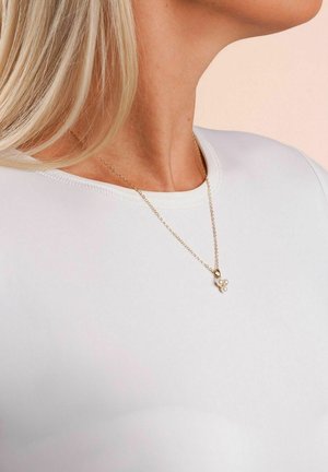 Nahaufnahme des Nackens einer Person, die ein weißes Hemd und eine goldene Kettenhalskette mit einem Anhänger aus drei kleinen Perlen trägt.