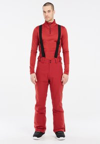 Rode skijas met hoge kraag en halve rits, gecombineerd met rode skibroek met zwarte bretels en een gladde textuur. Zwarte sneakers.