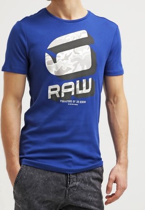 Homme portant une chemise à manches courtes bleue avec un grand logo "RAW" blanc et noir ainsi que le texte "PURVEYORS OF 3D DENIM" à l'avant.