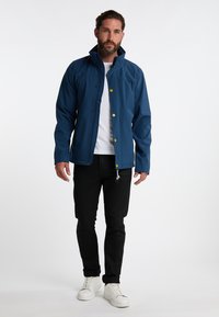 Kék softshell dzseki magas gallérral, patentgombokkal és zsebekkel. Fehér ing fölött viselve, fekete farmerrel és fehér cipővel párosítva.