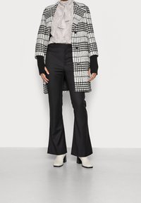 Czarny i biały płaszcz w kratę z szerokimi klapami, czarnymi prążkowanymi rękawami i falbankowym jasnym bluzką, w połączeniu z dopasowanymi czarnymi spodniami i białymi botkami do kostek.