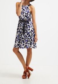 Robe bustier fleurie blanche, ornée de motifs bleus et noirs, taille cintrée avec une ceinture noire, associée à des sandales à plateforme marron.