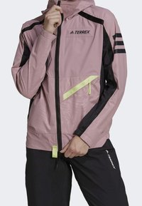 Rosa lättviktsjacka med svarta detaljer, med en dragkedja fram, huva och en reflekterande gul detalj på bröstet och fickan.