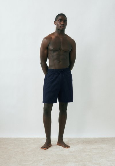 Hombre musculoso de pie descalzo sobre una alfombra, vistiendo pantalones cortos de color azul marino oscuro, con un fondo liso gris claro y las manos detrás de la espalda.