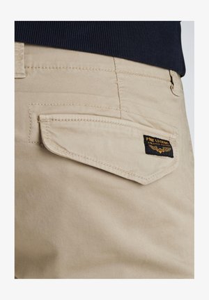 Beige látkové kalhoty se zadní kapsou s klopou, detailním šitím a černou etiketou se zlatým textem a designem s křídly.