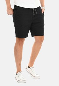 Schwarze Baumwollshorts mit elastischem Bund, Kordelzug und Seitentaschen. Kombiniert mit weißen Sneakern mit schwarzen Akzenten.